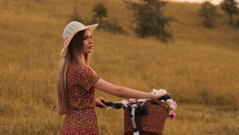 Rückansicht-In-Zeitlupe:-Eine-Schöne-Blondine-In-Einem-Kleid-Mit-Blumen-In-Einem-Korb-Und-Einem-Retro-Fahrrad-Geht-Auf-Der-Sommerwiese-Die-Straße-Entlang,-Schaut-Sich-Um-Und-Lächelt-Mit-Einem-Gefühl-Der-Freiheit.